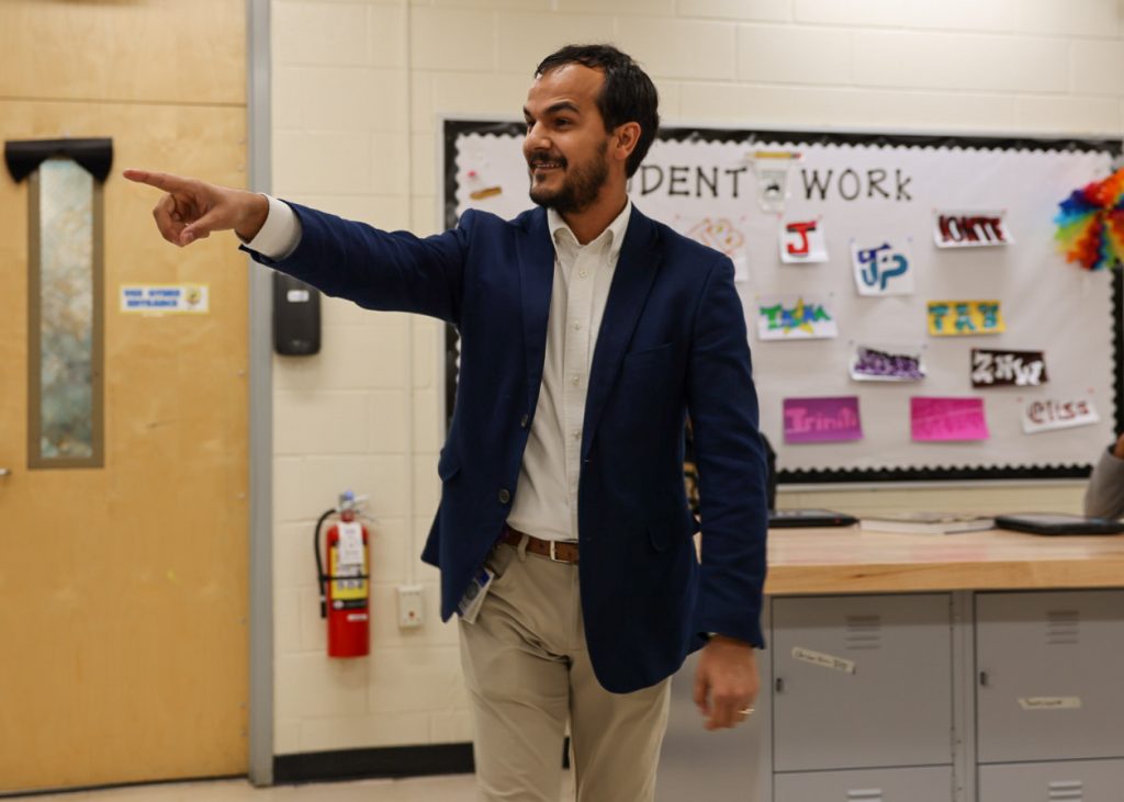 Assistant Solicitor Micah Chetta stands in a classroom, pointing toward an unseen student. A bulletin board is visible in the foreground.