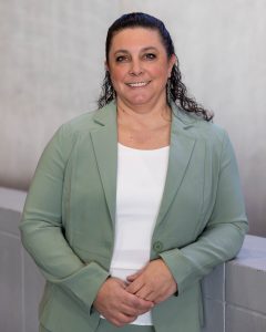 A professional portrait of a woman with dark, wavy hair pulled back, wearing a light green blazer over a white top. She is standing indoors, leaning slightly against a light-colored wall, and smiling warmly at the camera.